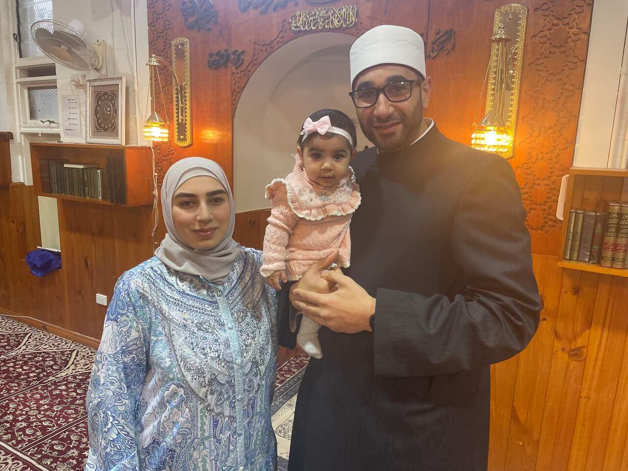 A woman in a hijab standing next to a man in a traditional Imam robe holding a baby girl.