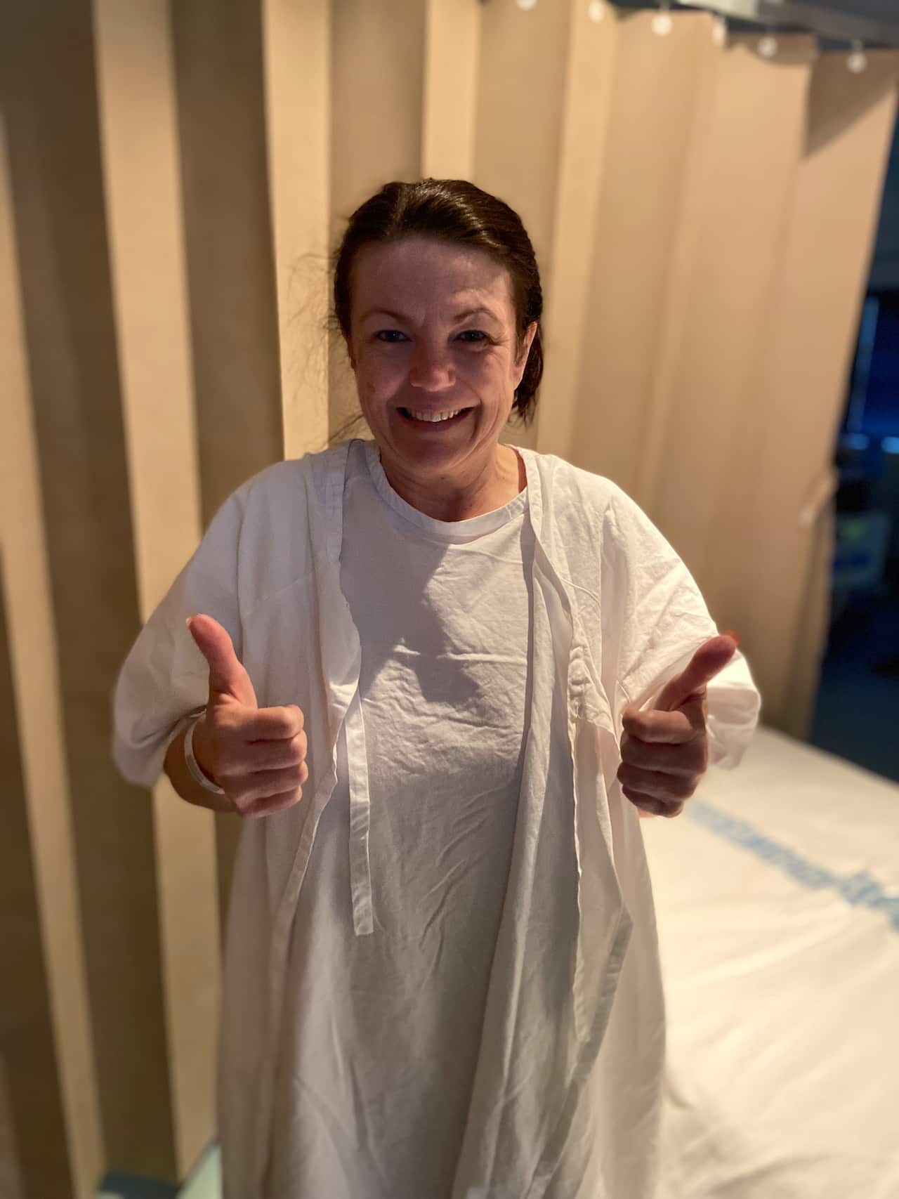 Woman standing in a hospital gown with her thumbs up.