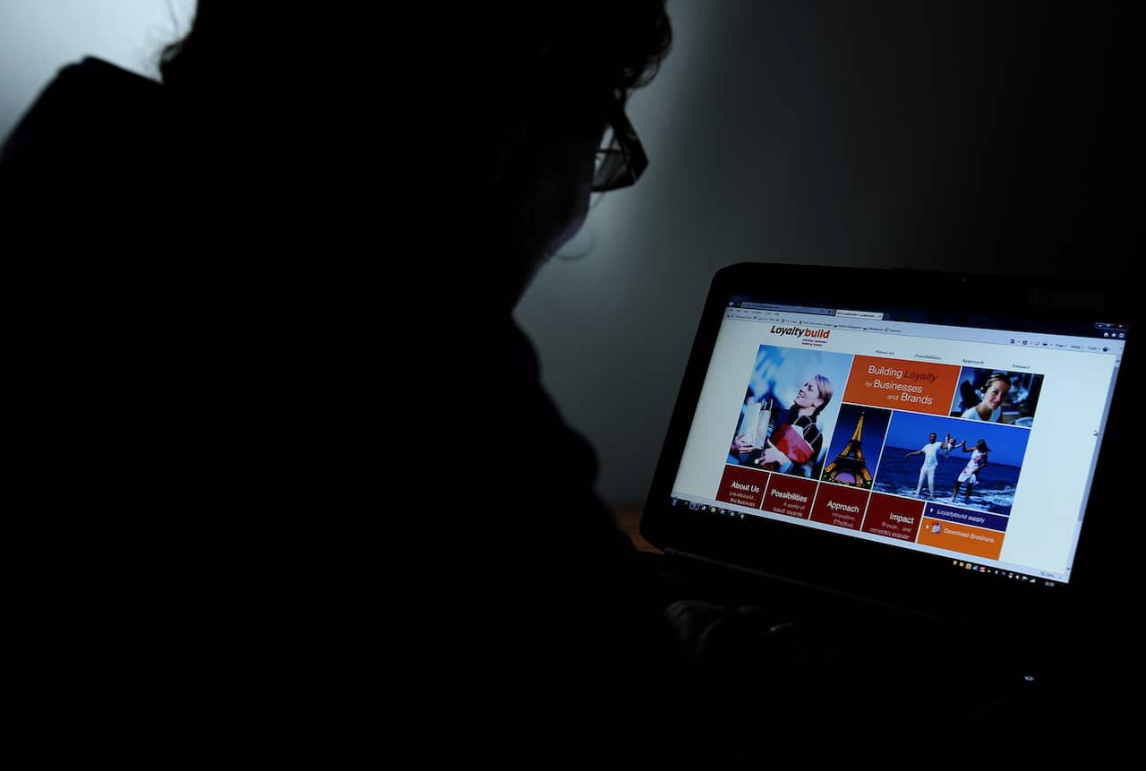A person on the left sits in a dark room reading a laptop that is on the right of frame.