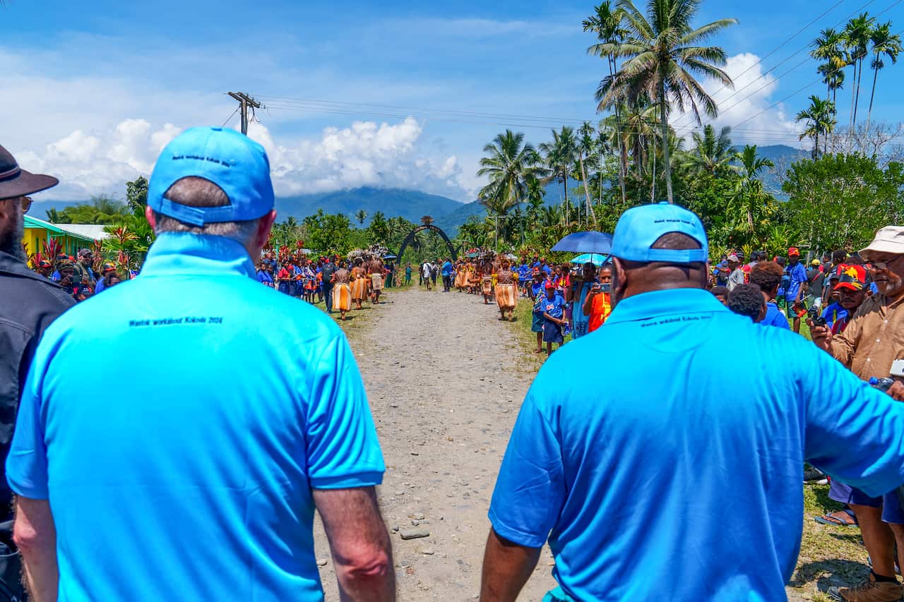 Rear view of Anthony Albanese (left) walking with James Marape on an unpaved road lined with people. Both men are wearing blue polo shirts and caps.