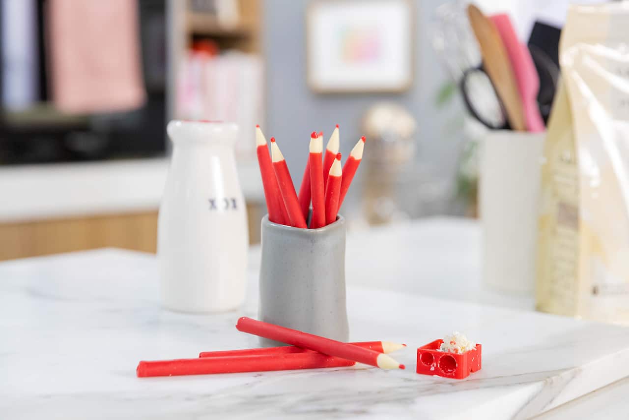 Seven red pencils stand in a tall grey pottery vessel. Three more lie on the table in front. 