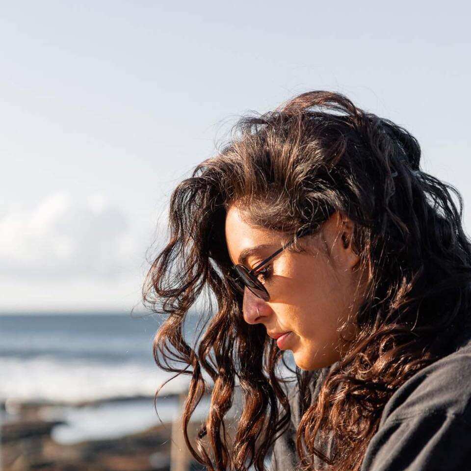 A woman with long, curly dark hair and sunglasses stares down at the ground on a beach