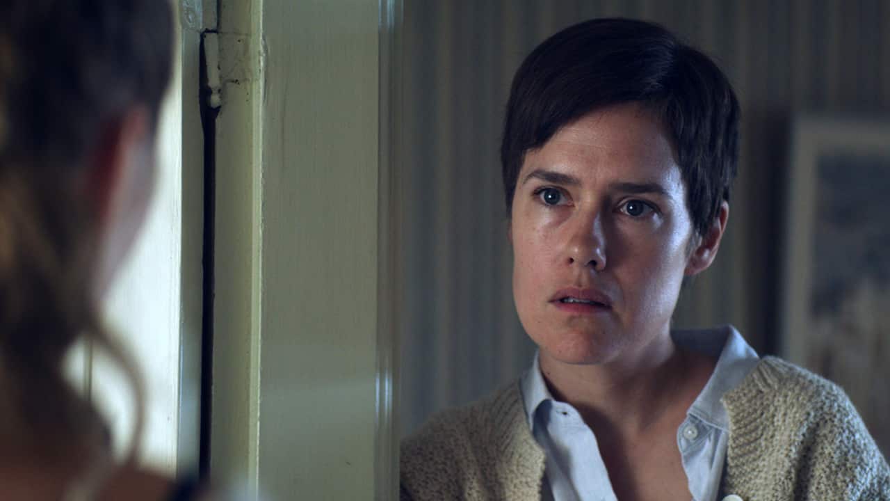 A woman stands in a doorway. The side of another woman can be seen, out of focus, at the edge of the image, facing her.  