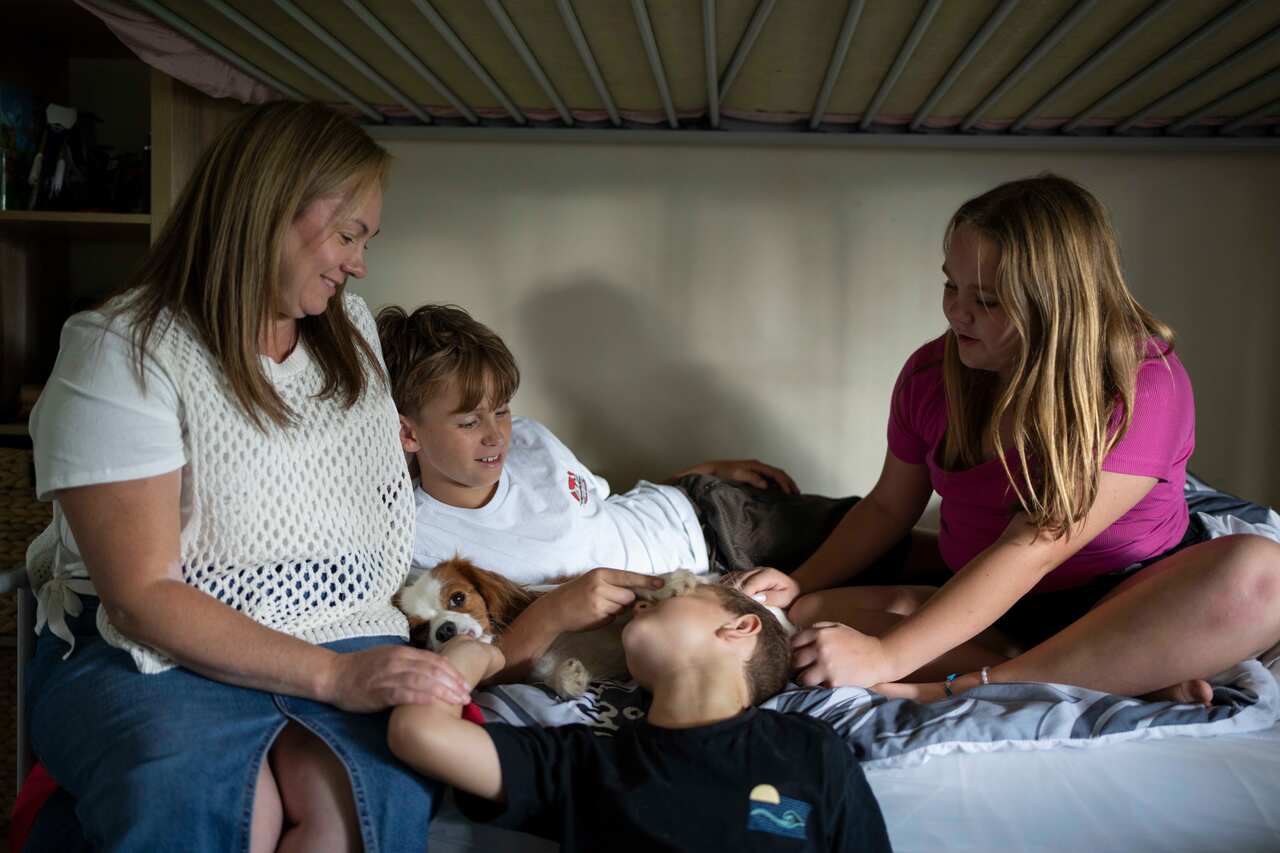 A woman with sandy hair sits on a bunk bed with three children: two young boys and a girl. 