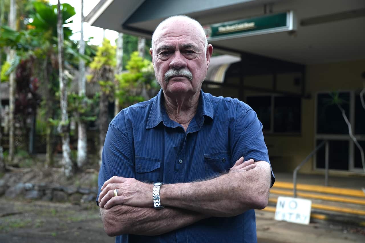 An older man in a blue shirt stands outside a building with his hands crossed in front.