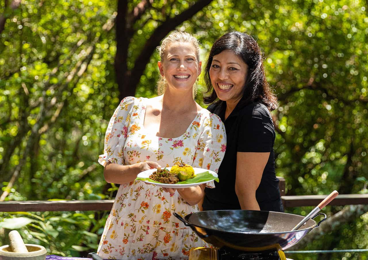 Two smiling women stand in front of a lush green plant-filled background. A wok sits in front of them. One woman holds a plate of beef rendang and a mound of yellow rice. 