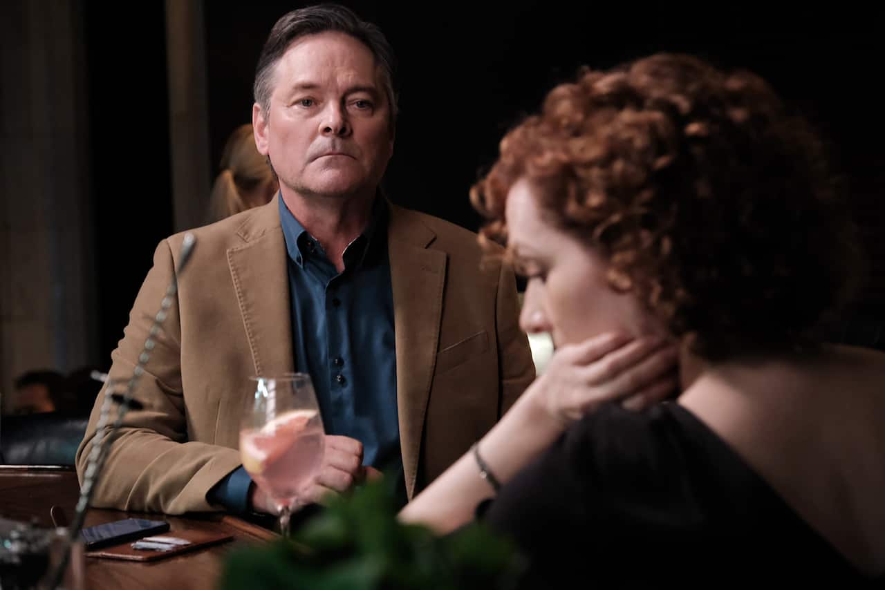 A man and a woman sit at a bar. She leans on her elbow, he faces her, 