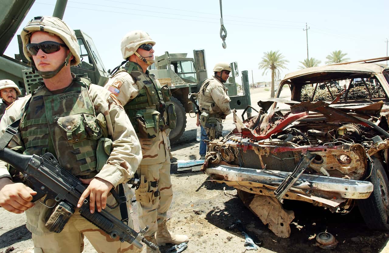 Soldiers with guns stand next to a a blown-up car. 