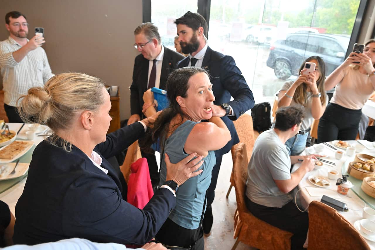 A woman reacts with surprise as others surround her, some capturing the moment with their phones in a restaurant.