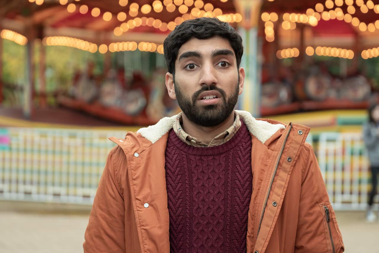 A bearded young man in a red knitted shirt and orange jacket stands in front of a brighly lit, colourful background.
