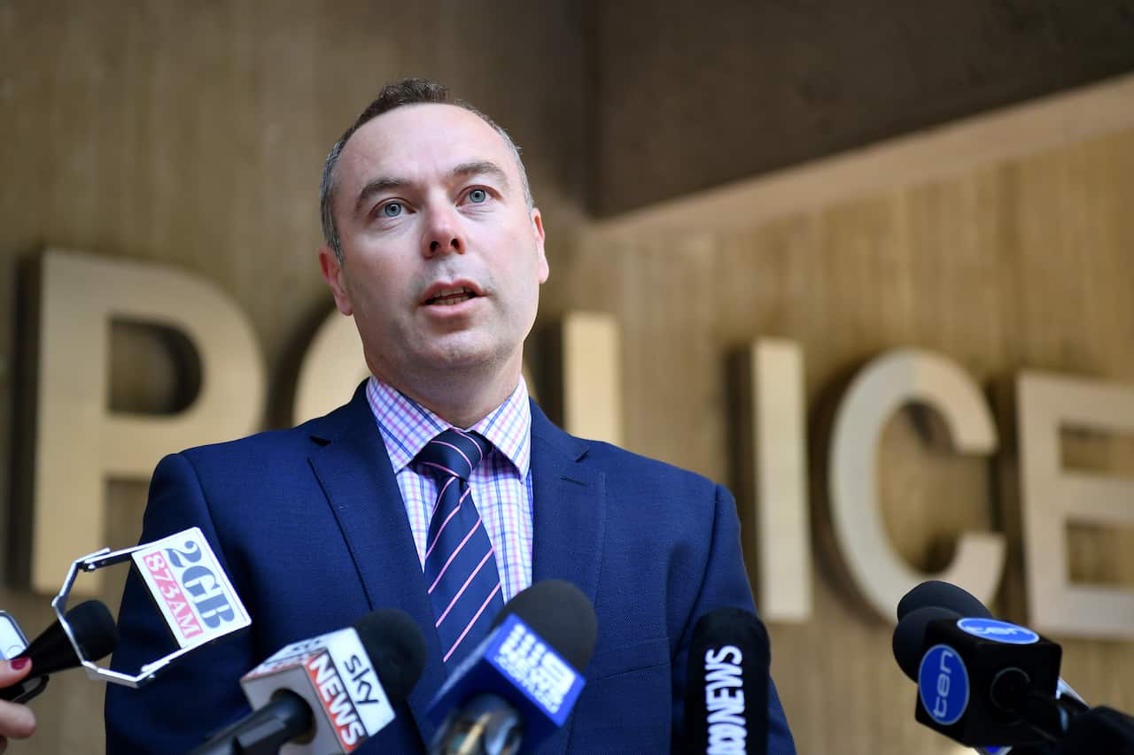 A man in a navy blue suit is talking into several microphones stationed below him, behind him is a sign that says 'POLICE'