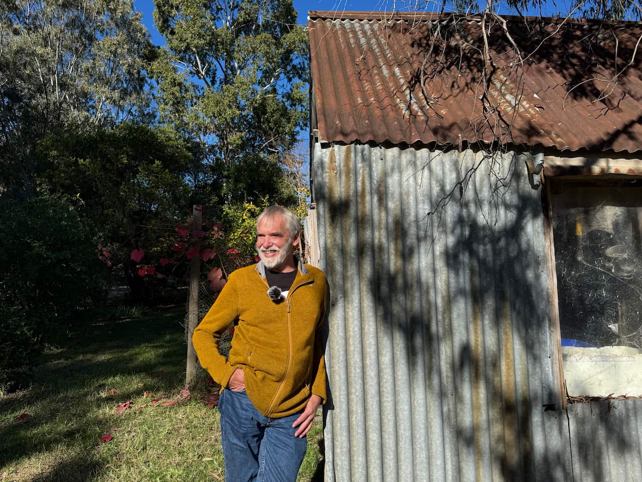 A man in a mustard zipper and blue jeans leans against the wall of a house with corrugated metal walls and a tin roof.