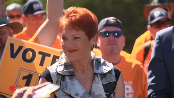 Pauline Hanson smiles while surrounded by supporters wearing orange shirts and holding campaign signs.
