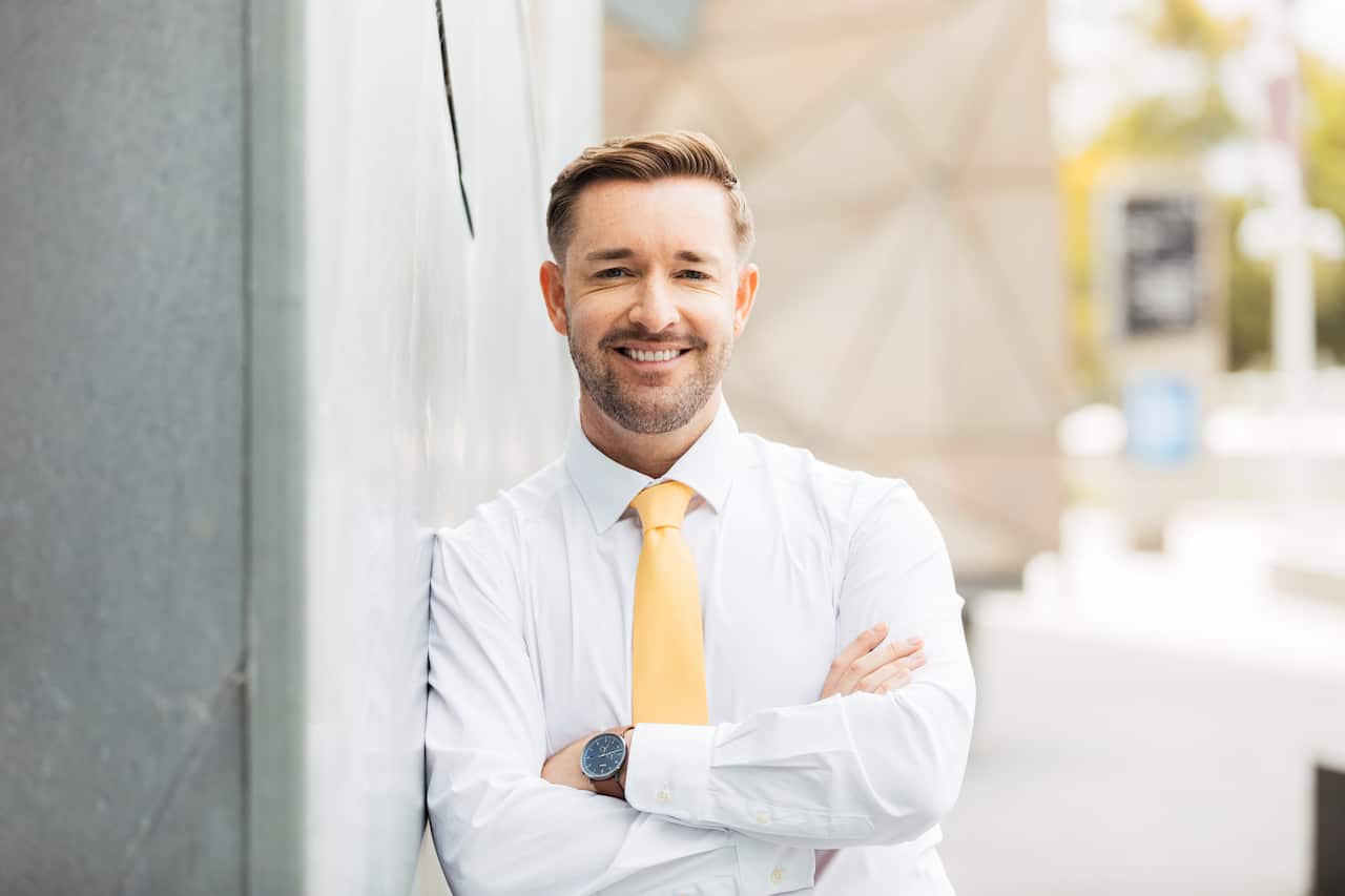A man with a beard is wearing a white shirt and yellow tie smiling with his arms crossed