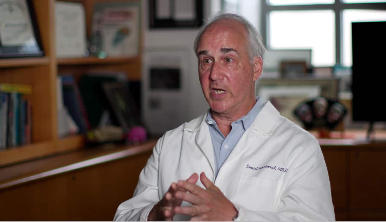 An older man in a white lab coat is speaking. His eyebrows are raised and his fingertips are pressed together. He is in mid-gesture. 