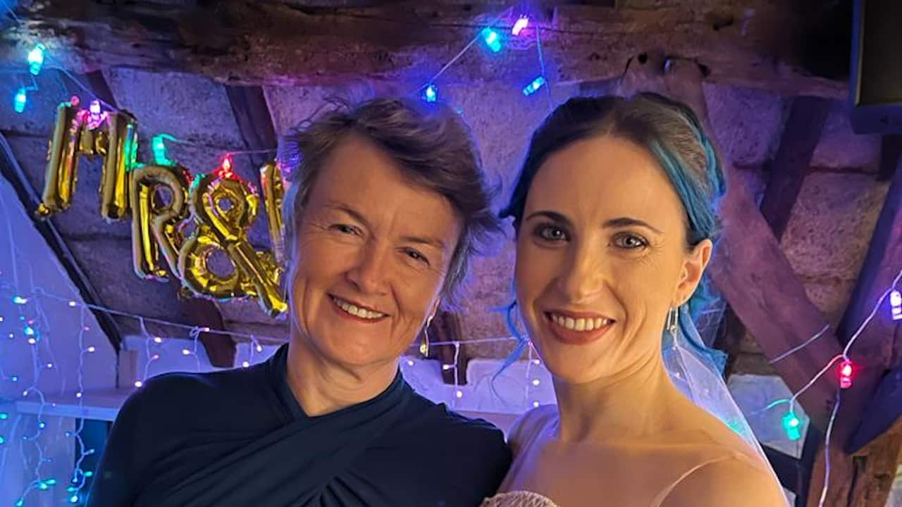 An older woman wearing a dark blue dress stands next to a younger woman in a wedding dress with dyed blue hair. They are both smiling.