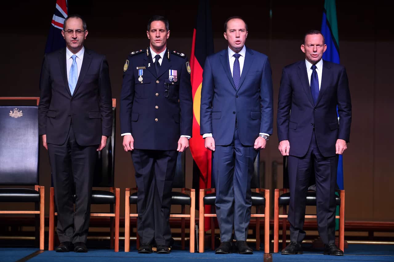 Four men in formal attire, one in a military cerimonial uniform, stand up singing the national anthem.