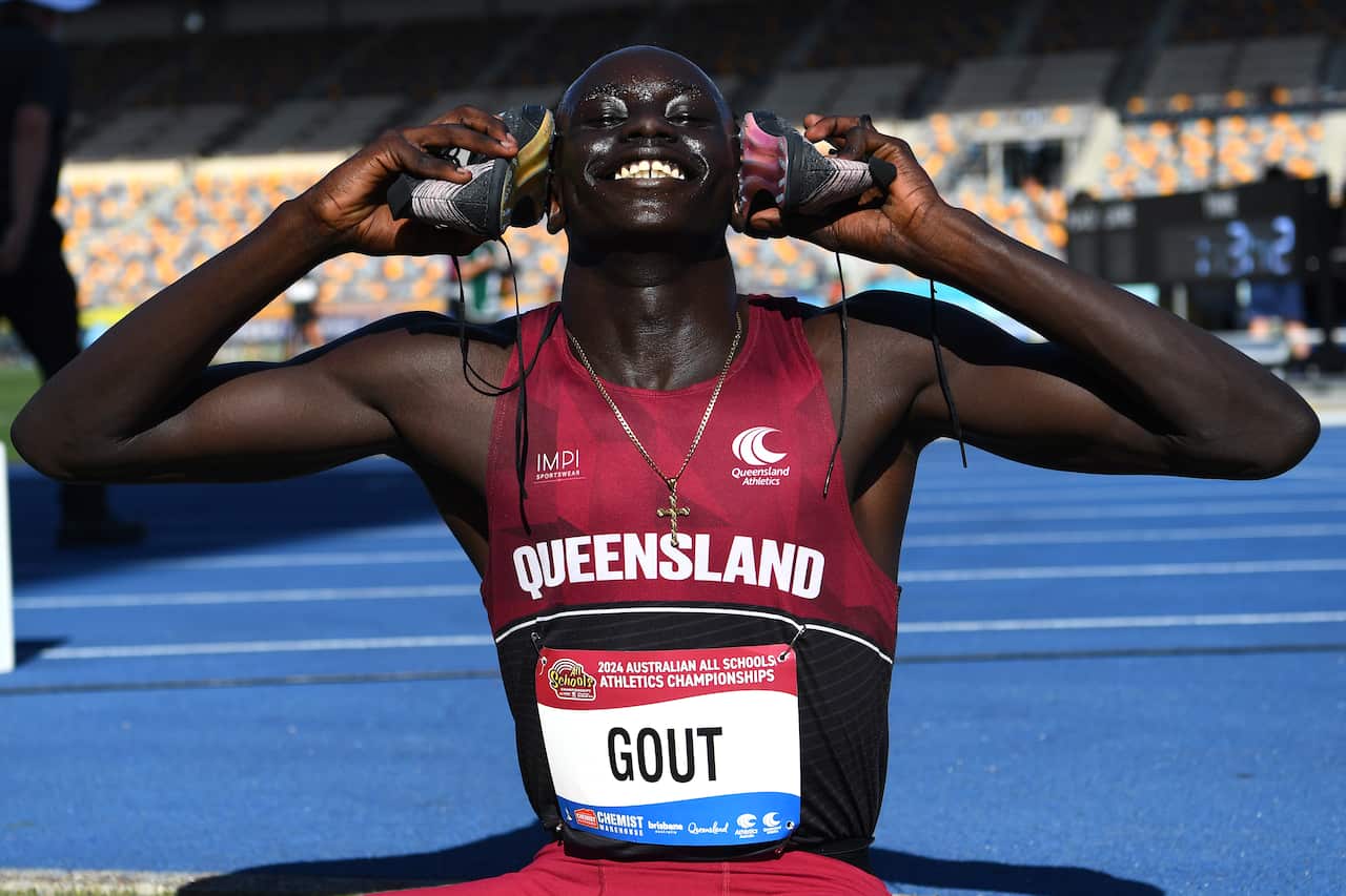 A young athlete wearing a red jersey with "Queensland" holds his running shoes up to his ears. He has a wide smile on his face. 