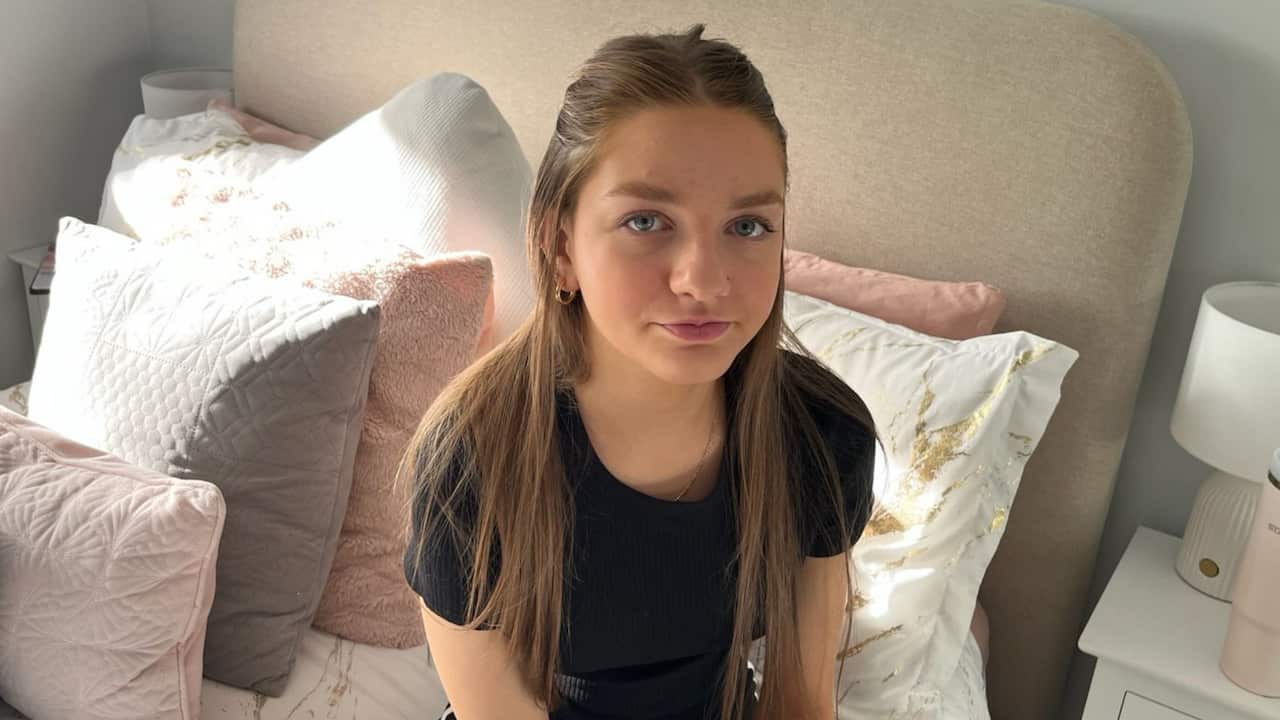 a teenage girl with long blonde hair looks neutrally sitting on her bed with white and pink pillows