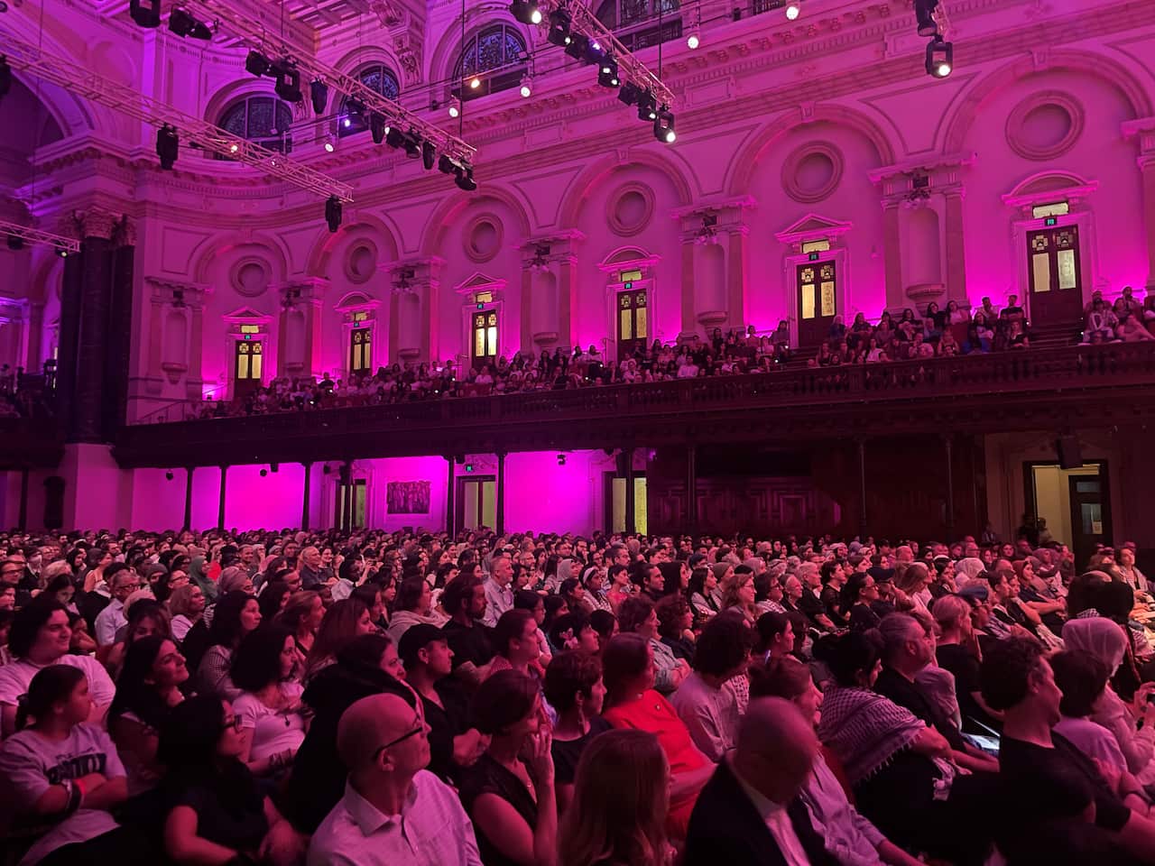 A hall is filled with rows of people sitting down. There is pink fluorescent lighting. 