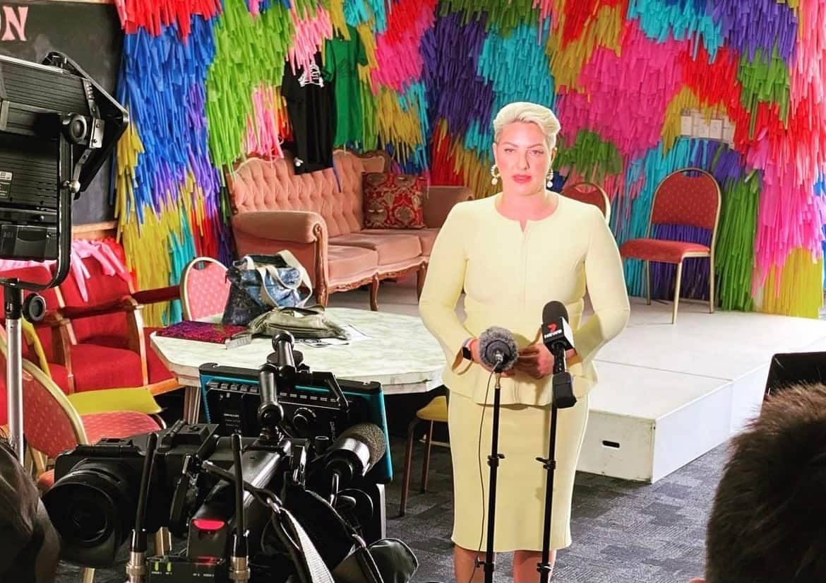 A woman in a yellow suit talks into a microphone. There is a television camera set up in front of her. 