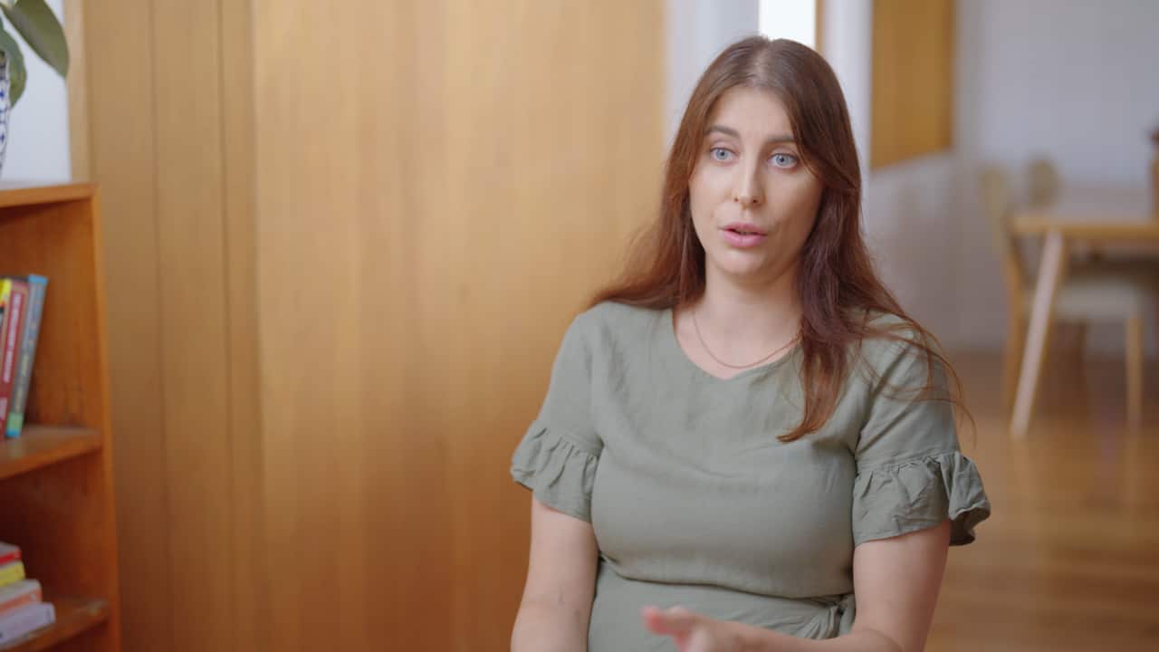 A woman with long, brown hair in a sage green top sits in a room with a bookshelf beside her and dining table behind her