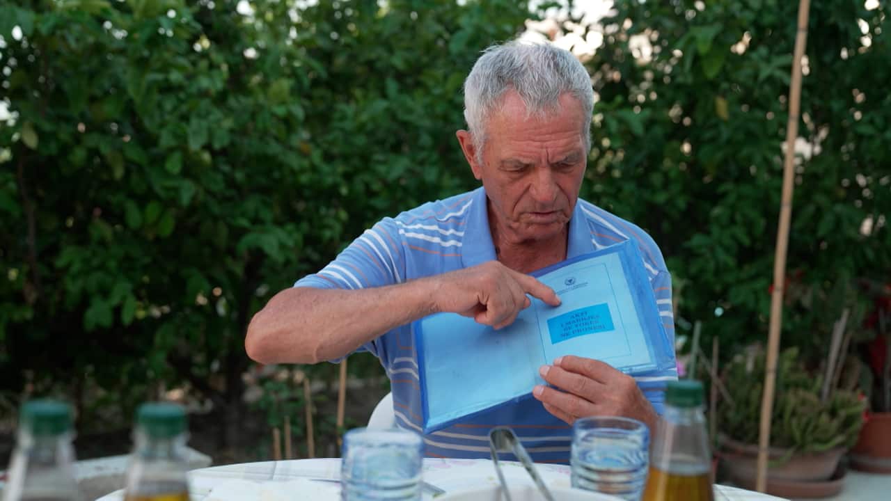 An older man in a blue polo shirt sits at an outdoor table. He holds a blue folder of documents and points to a page inside.