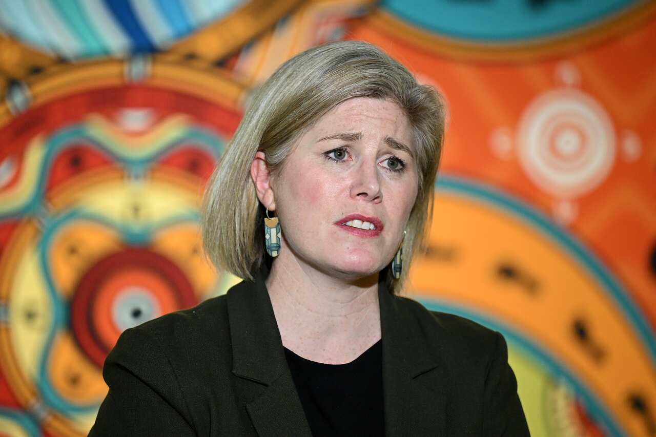 A woman in a black suit with blonde hair stands against a colourful background 