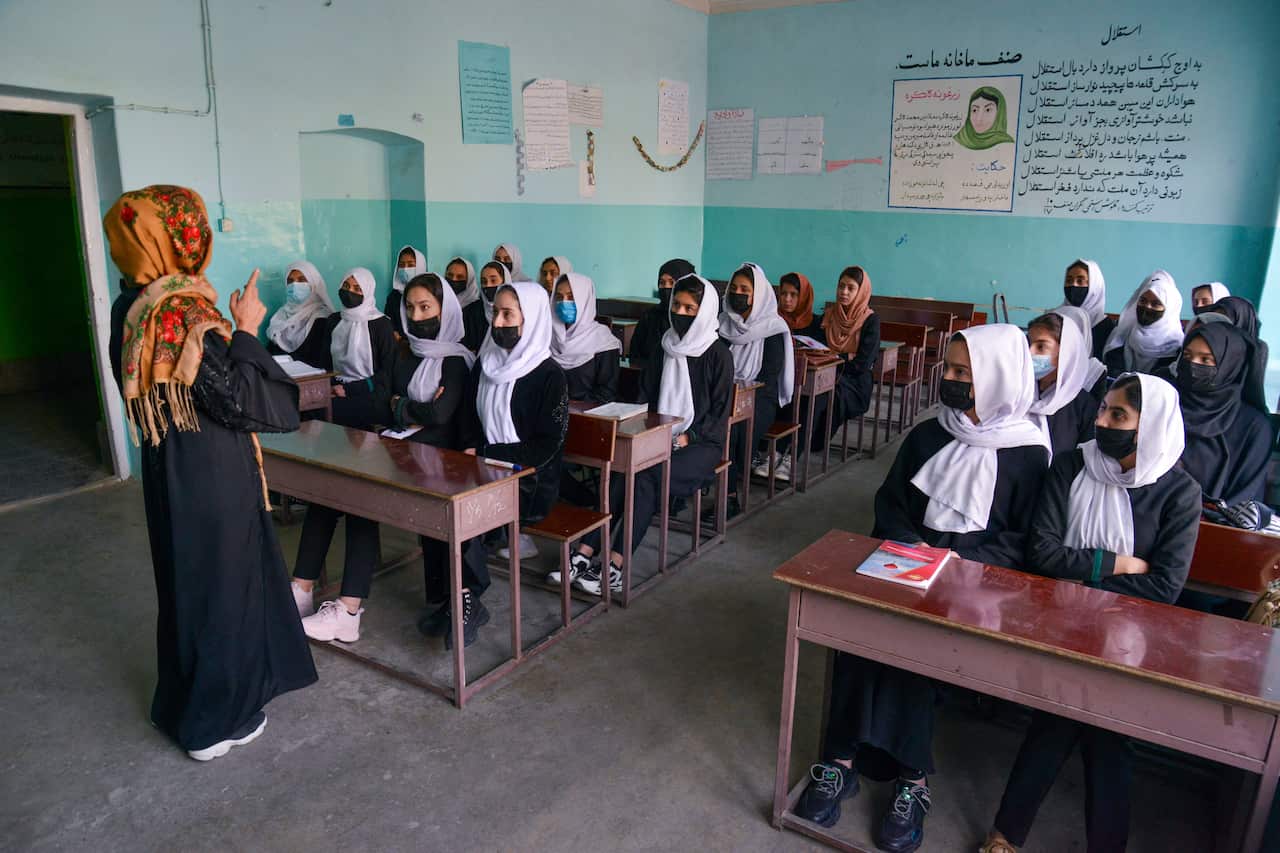 Girls are seen sitting at tables during a class while a teacher speaks.