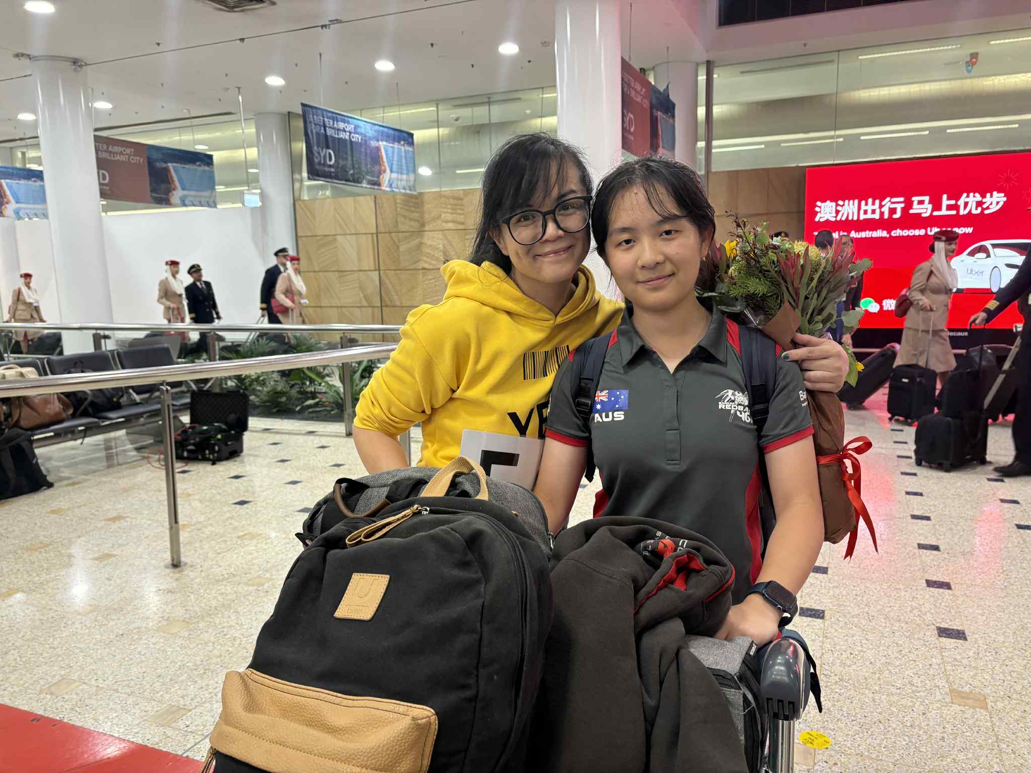 A middle aged Chinese women in a yellow hoodie holding a bunch of flowers with her arm around her daughter, a teenage girl wearing a dark grey shift with red trim, with an Australian flag on the breast, who is pushing a trolley of luggage.