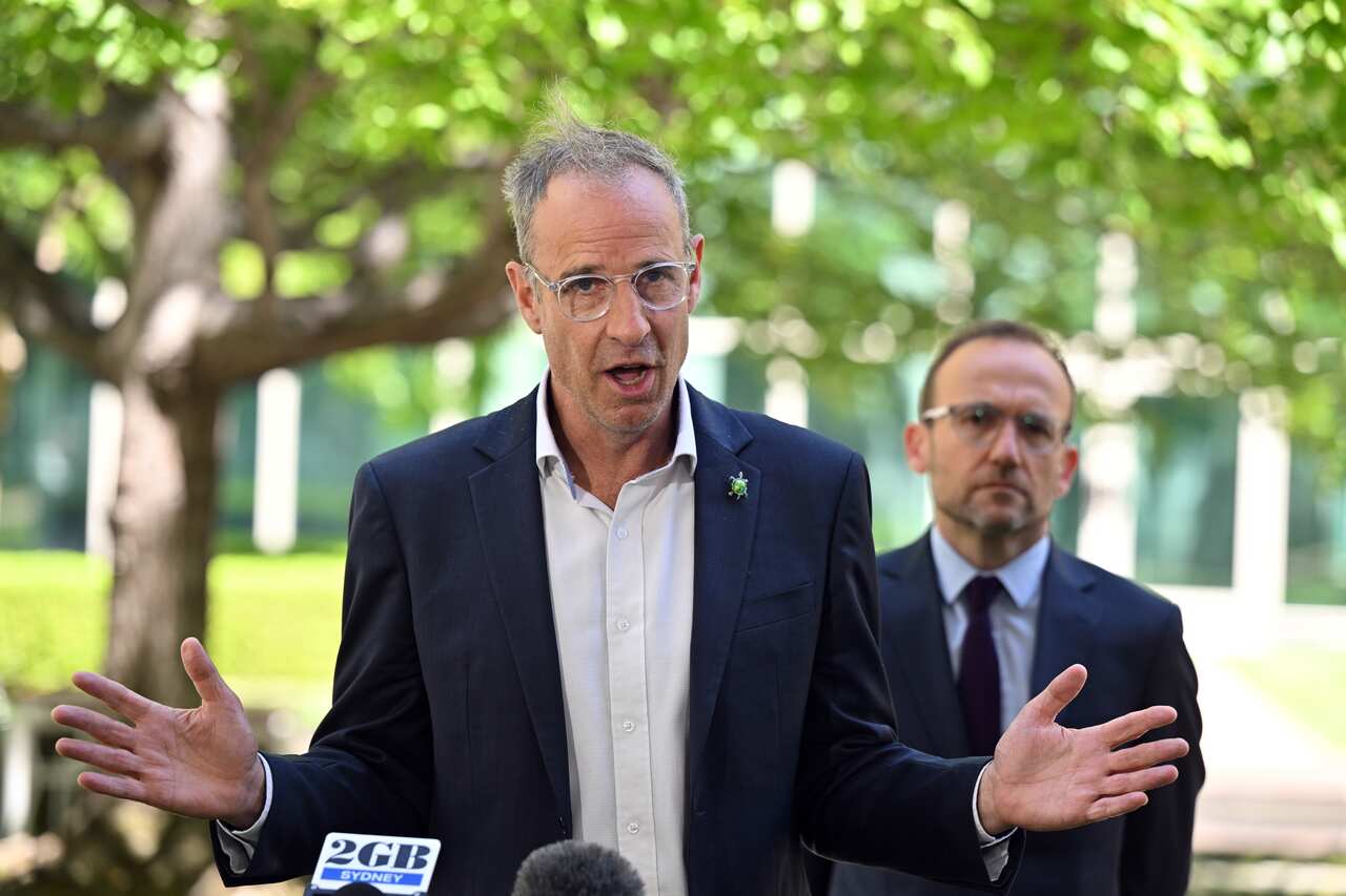 A man in a navy suit and white shirt speaking to press in front of a green park. Another man dressed similarly is behind him.