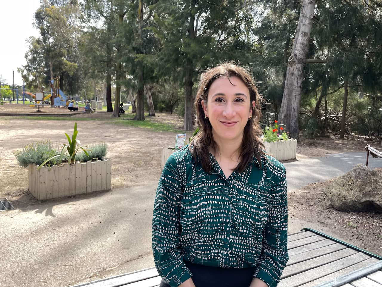 A woman in a green-and-white shirt standing in a park smiles.