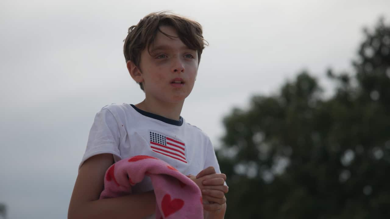 A young boy with smudges of dirt on his face looks into the distance. He is wearing a t-shirt with a US flag on it. 