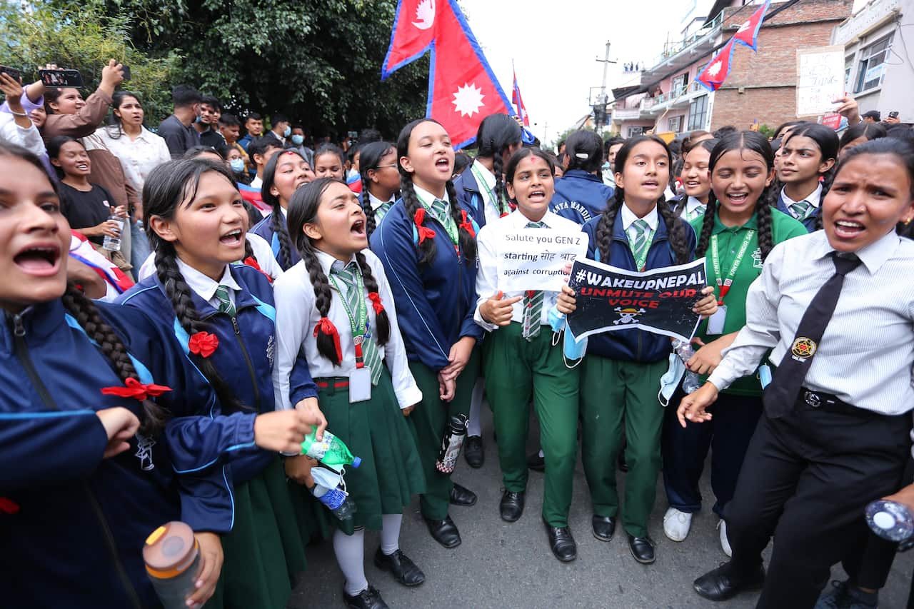A group of students in school uniforms is gathered on a street, with many of them shouting with their mouths open. Some students are holding protest signs.
