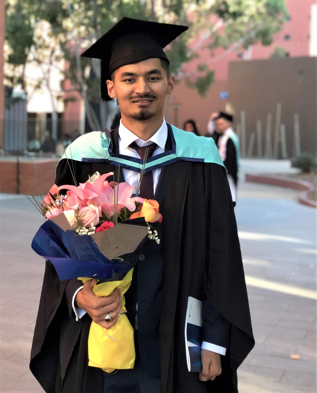 Kumail Jaffry holding a bunch of flowers in his graduation clothing.