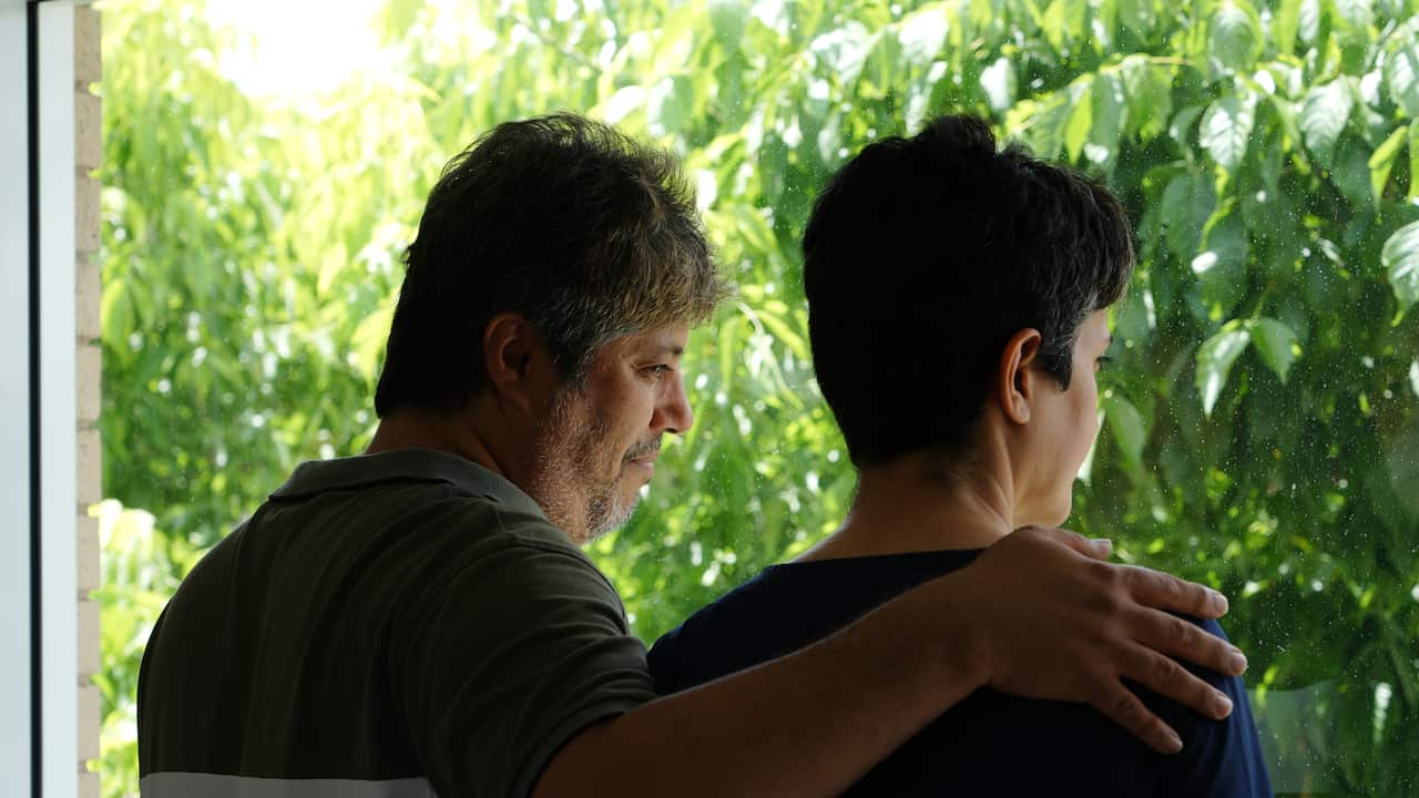 A middle-aged man of Iranian descent with his arm over the shoulder of a middle-aged woman with short dark hair. Both are looking pensively out the window, with the thick green leaves of a tree outside.
