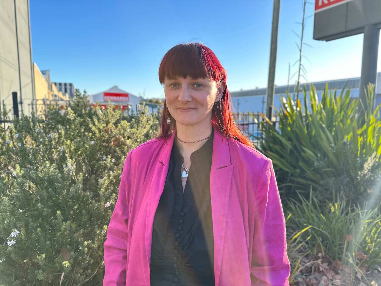 A woman wearing a pink blazer over a black shirt is standing in front of greenery.