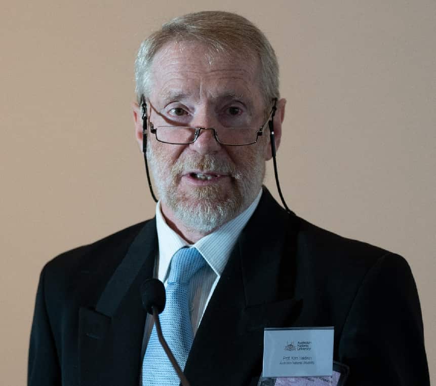 A grey-haired man in a black suit and blue tie with a name badge reading prof. Ken Baldwin