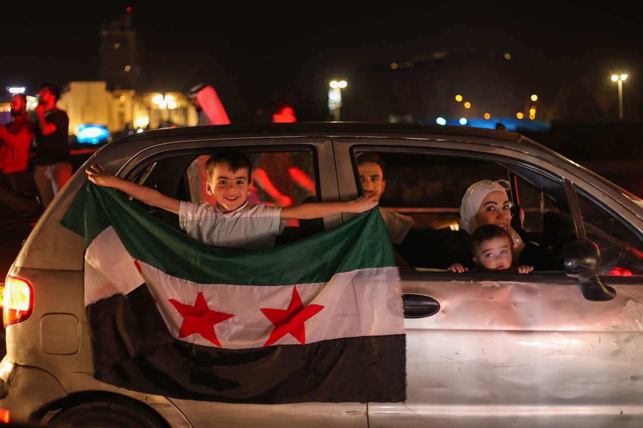 A child inside a car holds out a Syrian flag from the vehicle's window, and other passengers look on.