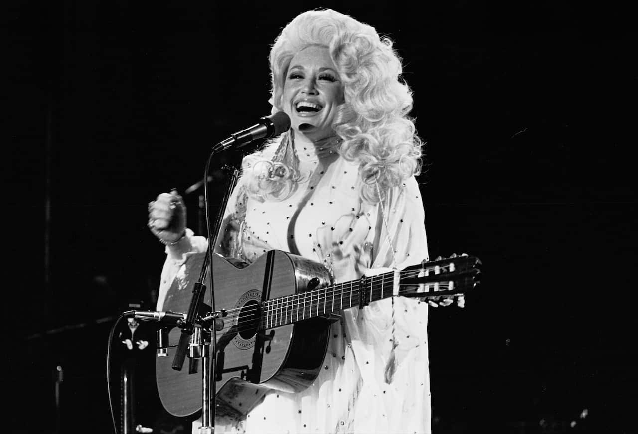 A smiling blonde woman wearing a white jumpsuit stands behind a microphone. She has a guitar on a strap across her body.