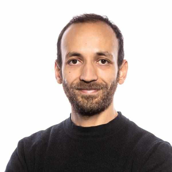 A man with dark hair and beard in a black t-shirt looks at the camera.