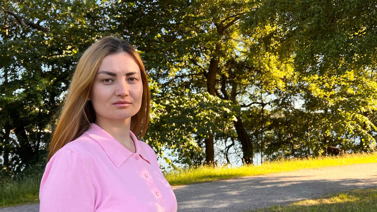 A woman with long blonde hair in a pink shirt looks straight into the camera. Green trees and a park path are visible in the background