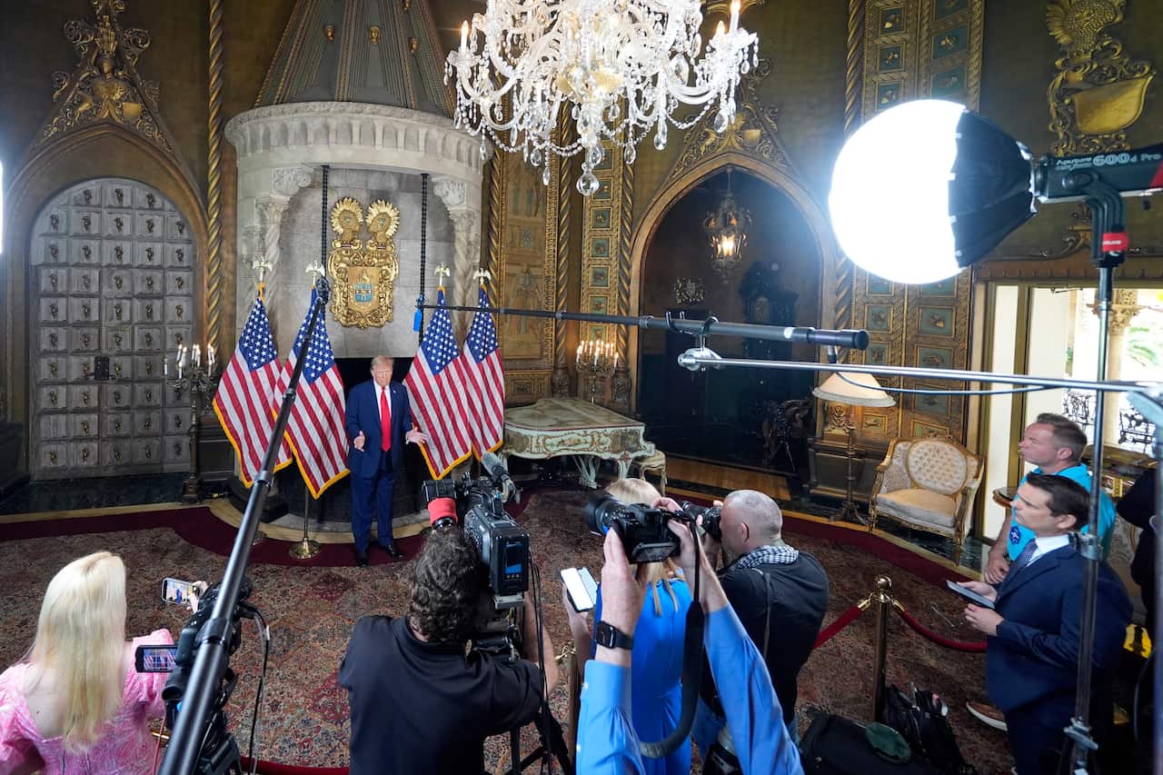 People with cameras and boom mics stand in front of a roped-off area where a man with a blue suit and red tie stands, delivering a speech in front of four American flags. 