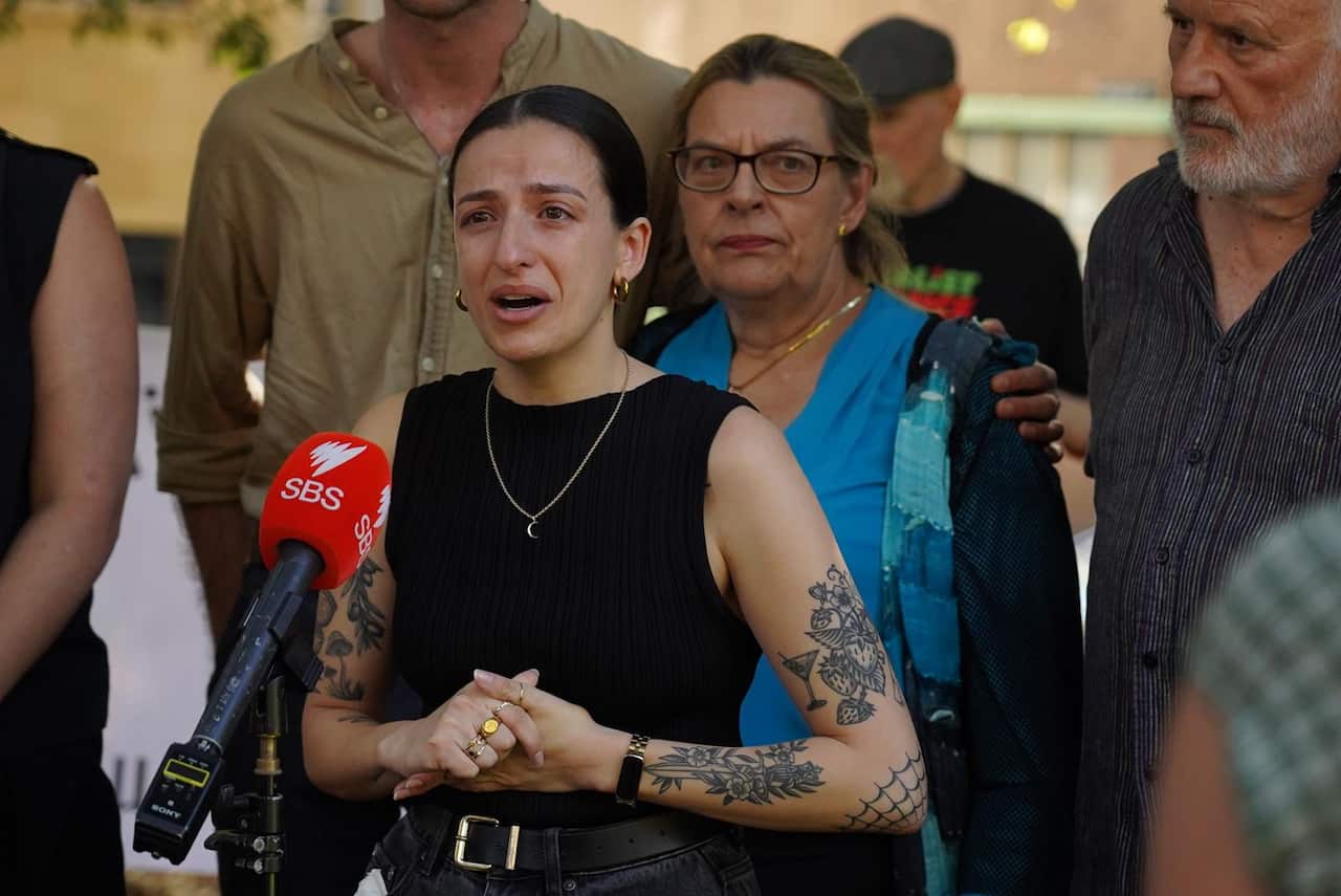A distressed-looking woman speaks into a microphone while others stand around her looking concerned.