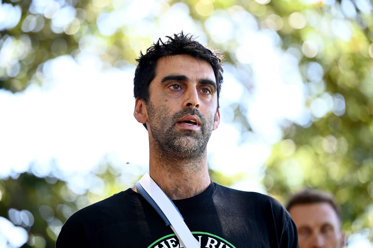 Surya McEwen, wearing a black shirt and speaking. There are trees behind him.