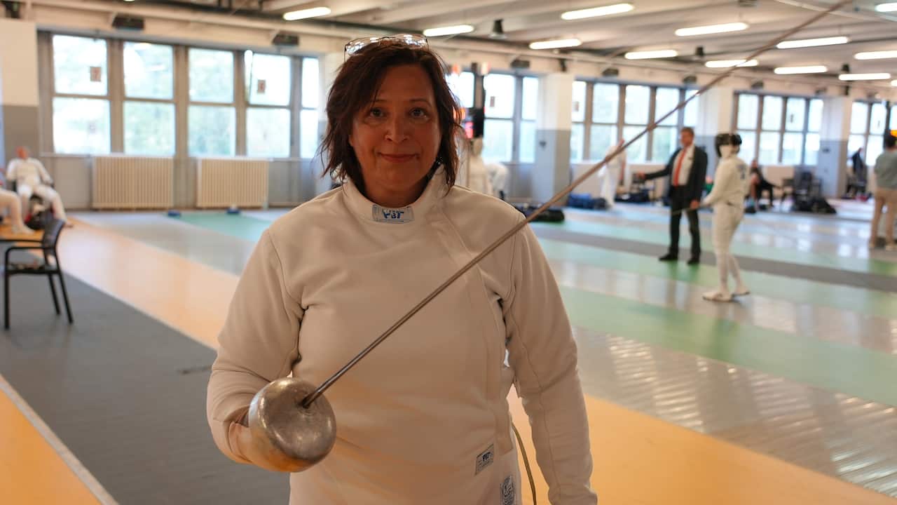 Una mujer blanca de mediana edad con cabello castaño, que lleva una espada de esgrima y un traje de esgrima blanco.