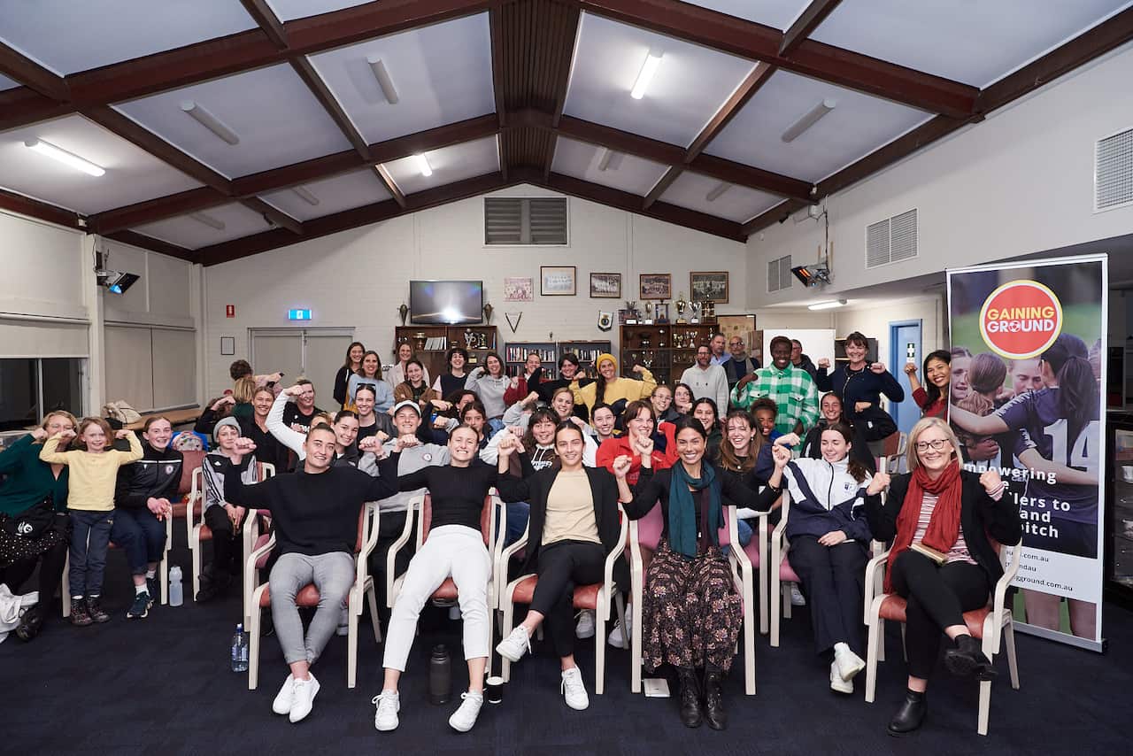 Women and girls are seated in chairs. They are holding their arms up in a gesture of strength. 