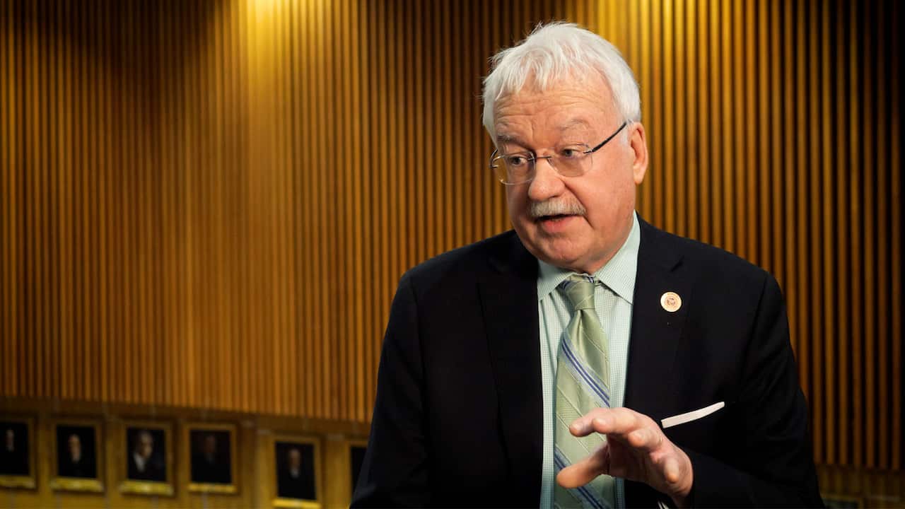 A grey-haired, moustachioed man wearing a dark suit and a light gree tie.