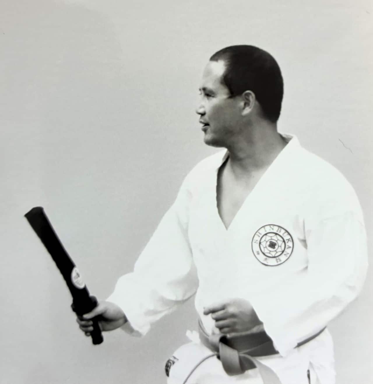 A man with black hair weaaring a white karate jacket holds a baton.