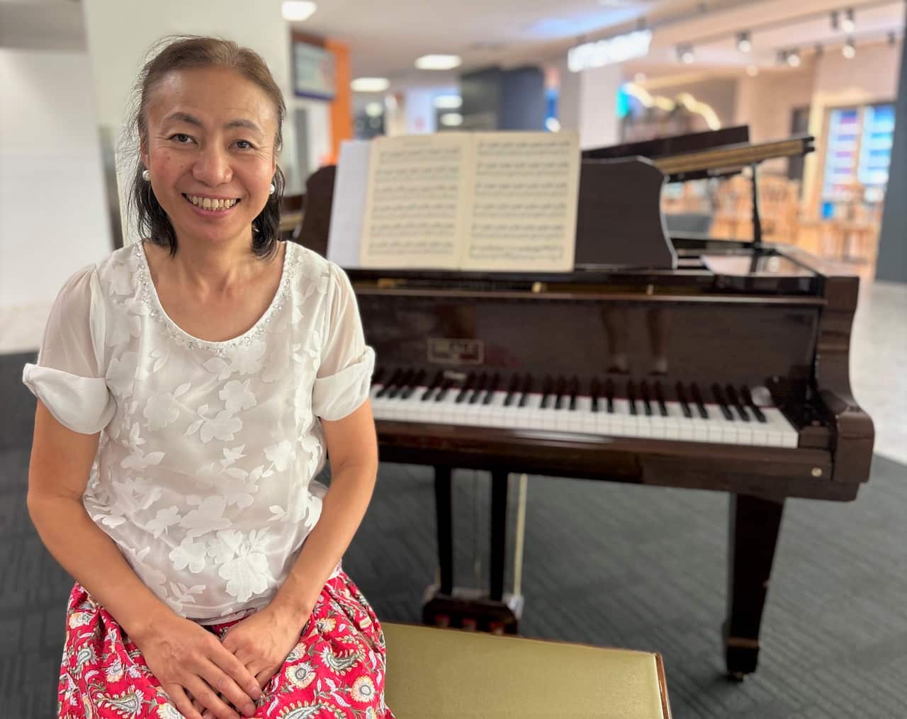 A woman sitting at a grand piano in a medical centre lobby smiling at the camera. 